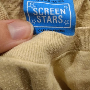 May include: Close-up of a tan t-shirt with a blue tag. The tag reads "MADE IN USA" and "SCREEN STARS" with a star design. The shirt is size S and made of cotton and polyester.
