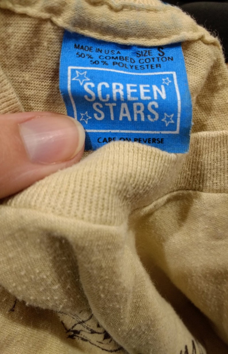 May include: Close-up of a tan t-shirt with a blue tag. The tag reads "MADE IN USA" and "SCREEN STARS" with a star design. The shirt is size S and made of cotton and polyester.