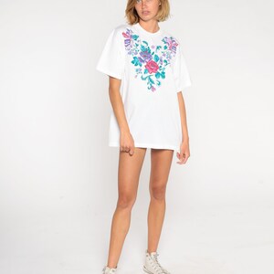 May include: White oversized t-shirt with a floral embroidery design on the front. The embroidery features pink, purple, and green flowers.
