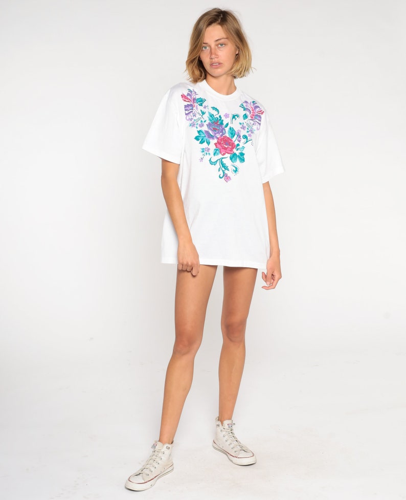 May include: White oversized t-shirt with a floral embroidery design on the front. The embroidery features pink, purple, and green flowers.