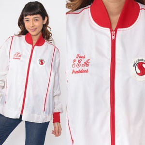 Peut inclure: Veste blanche à fermeture éclair avec des bordures rouges, un col et des poignets. La veste présente du texte brodé et un écusson circulaire sur la poitrine. Le texte indique "Toni SEA President" et "and a little bit more".