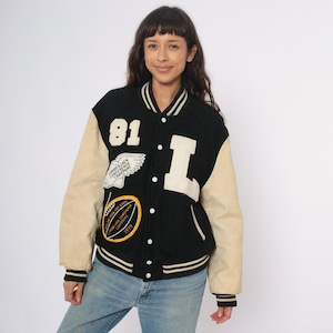 May include: Black and tan varsity jacket with white accents. Features the number "81" and a large "L" patch. Includes patches with text and a wing design. The jacket has a button-up front and ribbed cuffs.