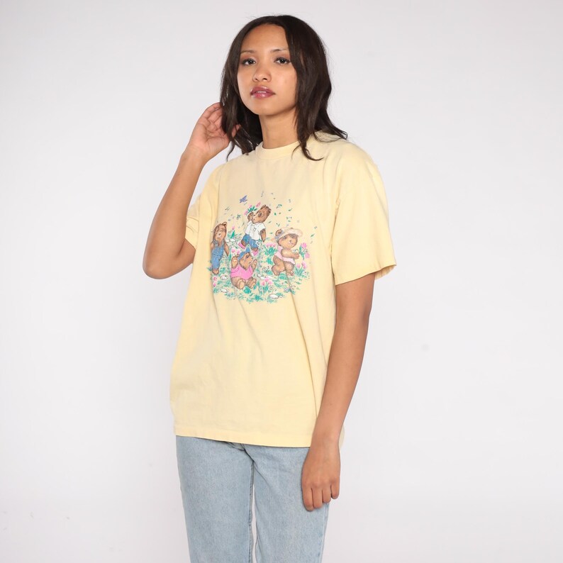 May include: A light yellow t-shirt featuring a graphic of cartoon teddy bears and children playing in a field of flowers. The t-shirt has a crew neck and short sleeves. The model is wearing light blue jeans.