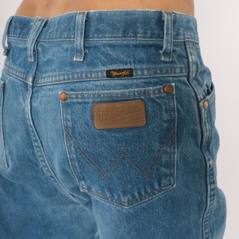 Puede incluir: Primer plano de la parte trasera de unos vaqueros azules. Los vaqueros presentan un parche de cuero marr&oacute;n con el logotipo "Wrangler" y una peque&ntilde;a etiqueta negra con el logotipo "Wrangler" encima del bolsillo trasero. Los vaqueros tienen remaches de color cobre.