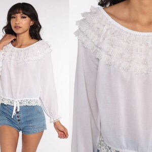 May include: White long-sleeved blouse with a ruffled neckline and lace trim. The blouse has a gathered waist and a slightly cropped length.