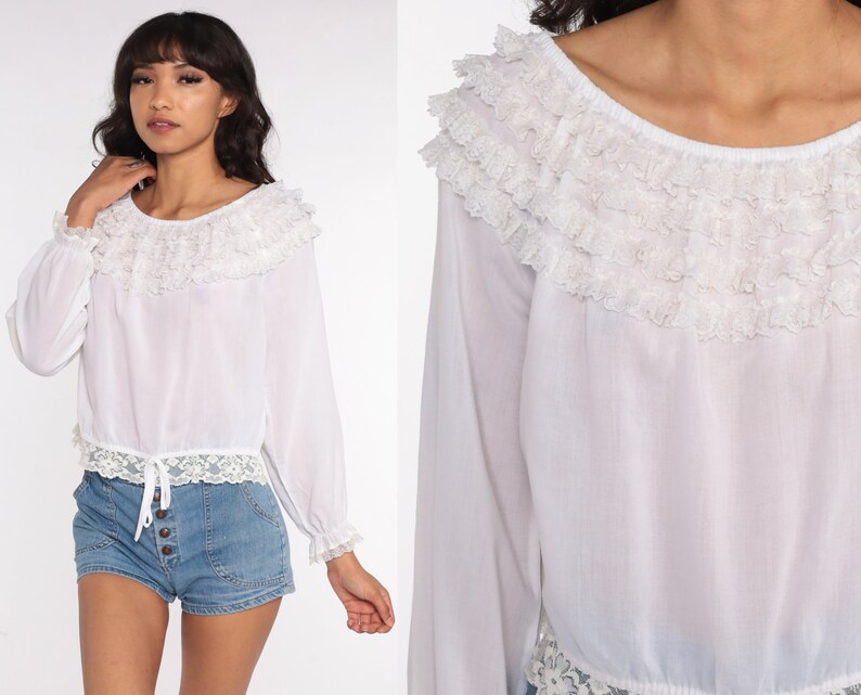 May include: White long-sleeved blouse with a ruffled neckline and lace trim. The blouse has a gathered waist and a slightly cropped length.