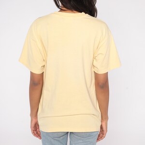 May include: A light yellow t-shirt with short sleeves. The shirt is made of a soft material and has a crew neck. The back of the shirt is visible, and the person wearing it is also wearing blue jeans.