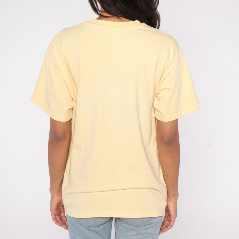 May include: A light yellow t-shirt with short sleeves. The shirt is made of a soft material and has a crew neck. The back of the shirt is visible, and the person wearing it is also wearing blue jeans.