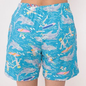 K&ouml;nnte beinhalten: Blaue Badeshorts mit einem Muster aus Haien und Surfern auf t&uuml;rkisfarbenen Wellen.