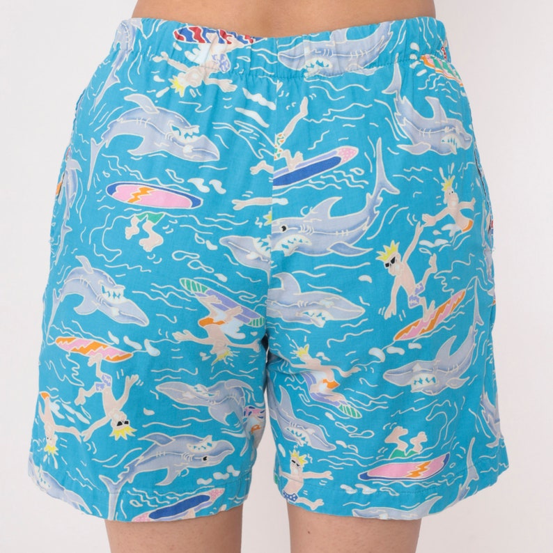 K&ouml;nnte beinhalten: Blaue Badeshorts mit einem Muster aus Haien und Surfern auf t&uuml;rkisfarbenen Wellen.