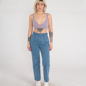 Op de afbeelding: Een vrouw draagt een lichtpaarse crop top en een blauwe high-waisted jeans. Ze staat met haar armen langs haar zijde en kijkt naar de camera.