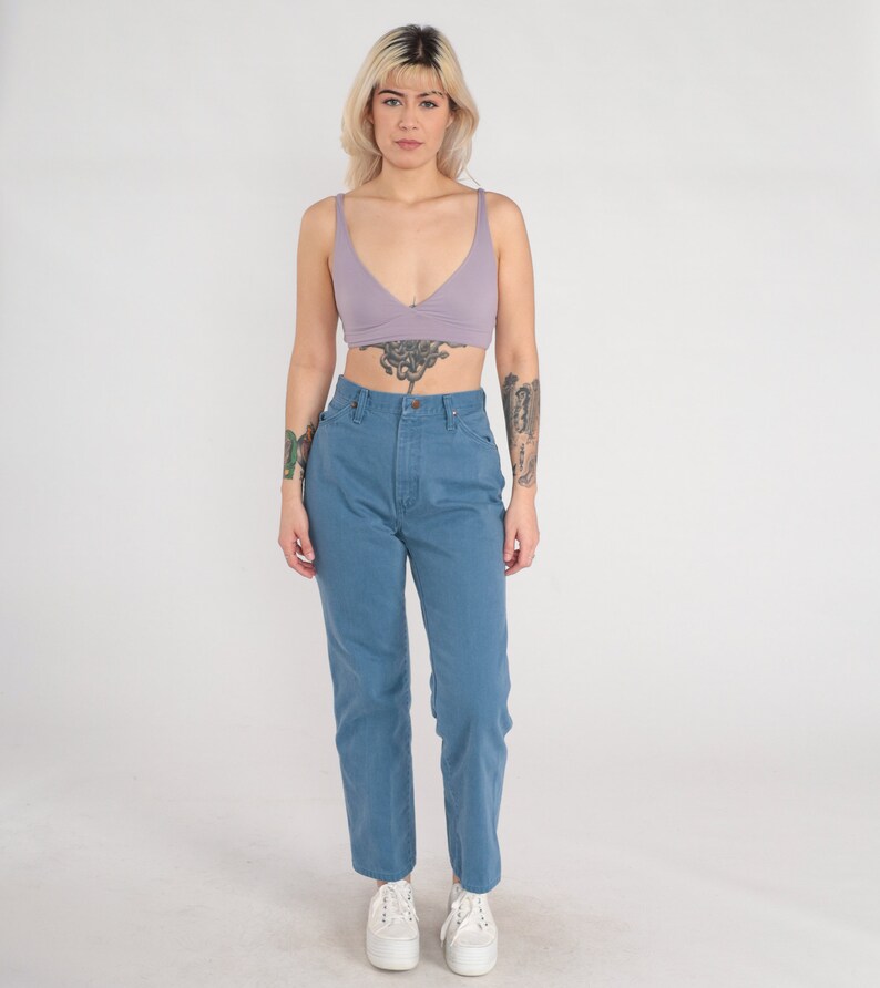 Op de afbeelding: Een vrouw draagt een lichtpaarse crop top en een blauwe high-waisted jeans. Ze staat met haar armen langs haar zijde en kijkt naar de camera.