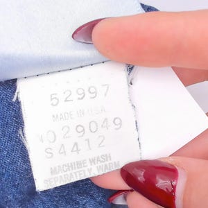 Puede incluir: Una etiqueta de cuidado de tela blanca unida a una prenda azul. La etiqueta presenta el texto 52997, MADE IN USA, NO 29049, S 4412 y MACHINE WASH SEPARATELY, WARM. La etiqueta es rectangular con texto negro impreso sobre un fondo blanco.