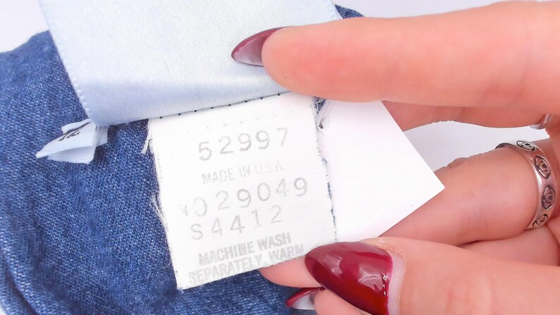 Puede incluir: Una etiqueta de cuidado de tela blanca unida a una prenda azul. La etiqueta presenta el texto 52997, MADE IN USA, NO 29049, S 4412 y MACHINE WASH SEPARATELY, WARM. La etiqueta es rectangular con texto negro impreso sobre un fondo blanco.