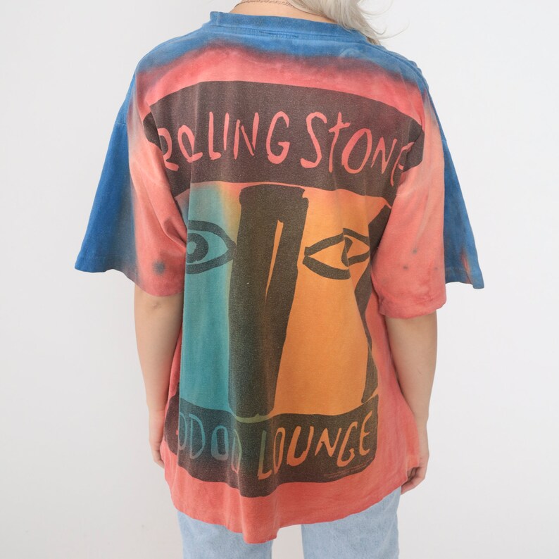 May include: A tie-dye t-shirt with a vintage Rolling Stones design. The shirt is red, orange, blue, and green with a black graphic of a face and the text "Rolling Stone" and "Odd Lounge".