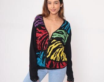 May include: A black cardigan with a V-neck and long sleeves. The front features a vibrant zebra stripe pattern in rainbow colors, embellished with sequins. The sleeves and trim have sequin details. The cardigan is worn with blue jeans.