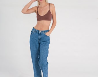 May include: A brown bra with thin straps and high-waisted blue jeans. The jeans have a classic five-pocket design. The model is wearing brown suede sandals. The background is plain white.