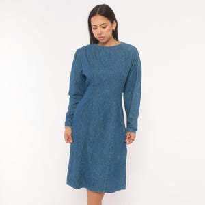 Puede incluir: Un vestido de mezclilla azul de tono medio con mangas largas, cuello redondo y detalles de costuras verticales en la parte delantera del corpi&ntilde;o. El vestido tiene una cintura entallada y una falda hasta la rodilla, hecho de tela de algod&oacute;n.