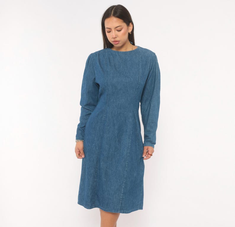 Puede incluir: Un vestido de mezclilla azul de tono medio con mangas largas, cuello redondo y detalles de costuras verticales en la parte delantera del corpi&ntilde;o. El vestido tiene una cintura entallada y una falda hasta la rodilla, hecho de tela de algod&oacute;n.