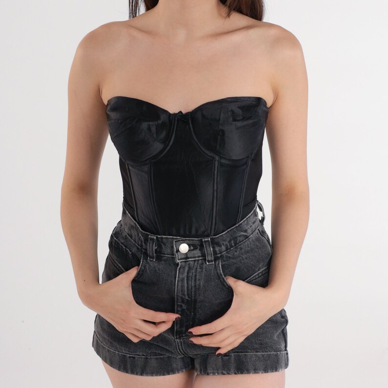 Puede incluir: Un cors&eacute; negro de sat&eacute;n sin tirantes con varillas y escote de coraz&oacute;n. El cors&eacute; se combina con pantalones cortos de mezclilla oscura.