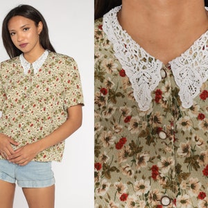 K&ouml;nnte beinhalten: Eine kurz&auml;rmlige Blumenbluse mit wei&szlig;em Spitzenkragen. Die Bluse hat eine Knopfleiste und ein Muster aus kleinen roten und wei&szlig;en Blumen auf hellgr&uuml;nem Hintergrund. Der Spitzenkragen ist ein auff&auml;lliges Detail.