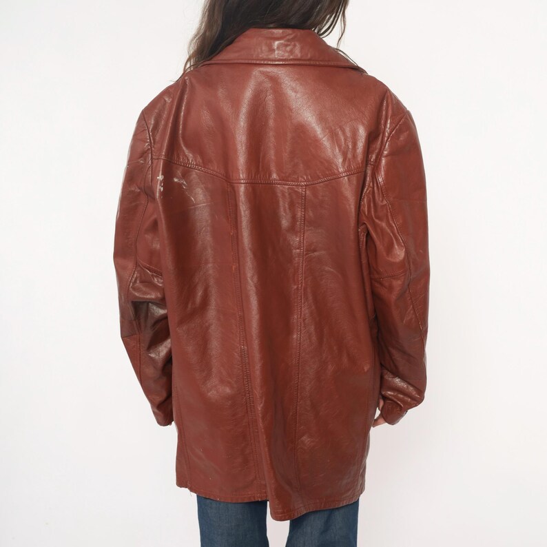 May include: A vintage brown leather jacket with a collared neckline and button closure. The jacket has a longline silhouette and a slightly worn appearance, suggesting it's a pre-owned item. The jacket is worn by a person wearing blue jeans.