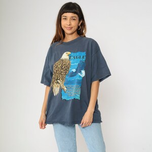 May include: A dark blue t-shirt featuring a graphic of a bald eagle on a blue and white background. The text "Bald Eagle" is printed on the shirt.