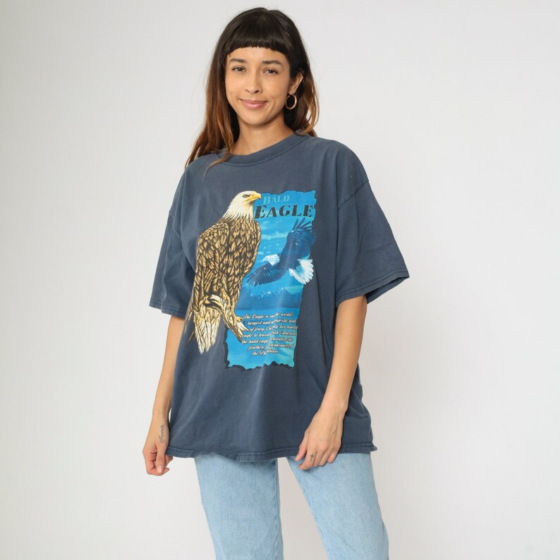May include: A dark blue t-shirt featuring a graphic of a bald eagle on a blue and white background. The text "Bald Eagle" is printed on the shirt.