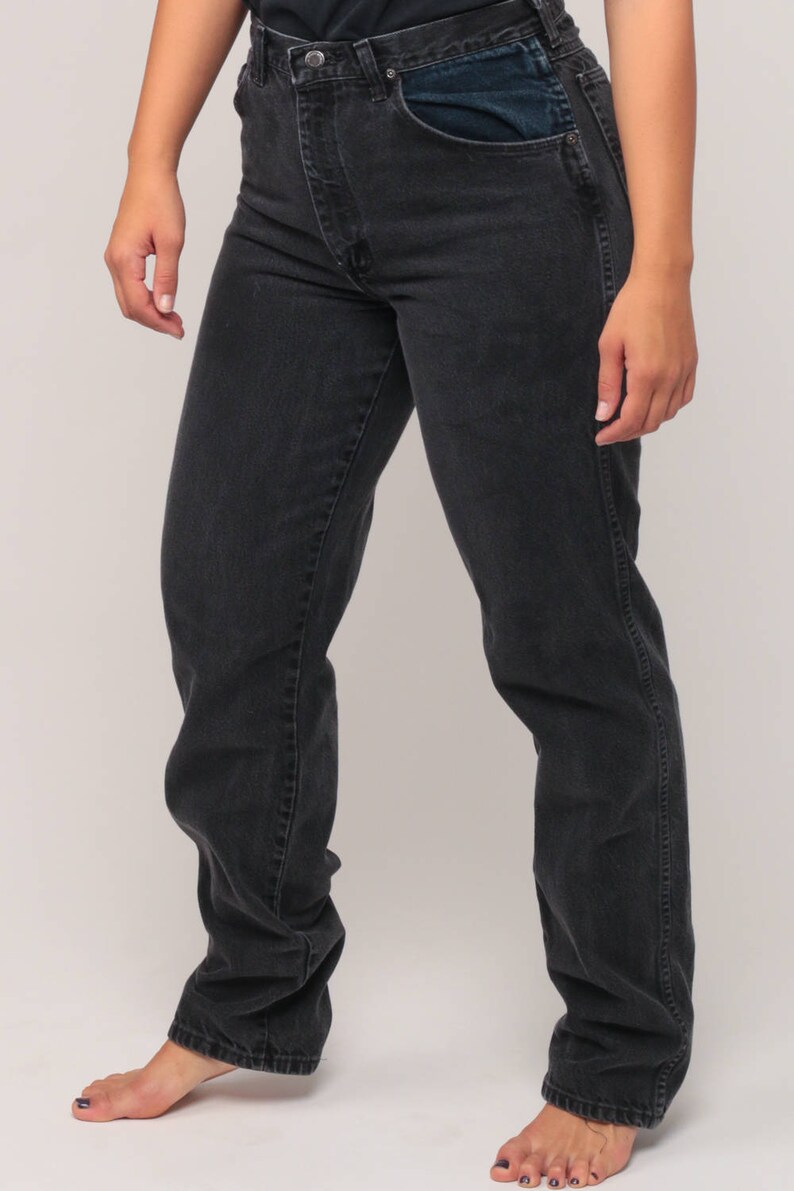 May include: Dark gray denim jeans with a classic five-pocket design. The jeans have a straight leg fit and a button and zipper closure. The front pockets have a contrasting dark blue fabric. The model is wearing the jeans.