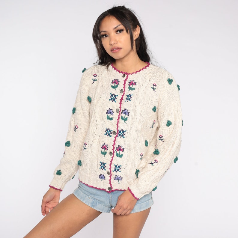 May include: A cream-colored knitted cardigan with a floral embroidered design. The cardigan has a button-up front and a crocheted trim. The cardigan has a textured knit and is decorated with small green pom-poms.