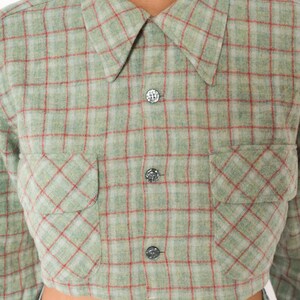 Puede incluir: Una camisa corta abotonada con un patr&oacute;n de cuadros en verde salvia y blanco con finas l&iacute;neas rojas. La prenda presenta un cuello en punta, mangas largas y dos bolsillos delanteros en el pecho con solapas, asegurados por tres botones oscuros.
