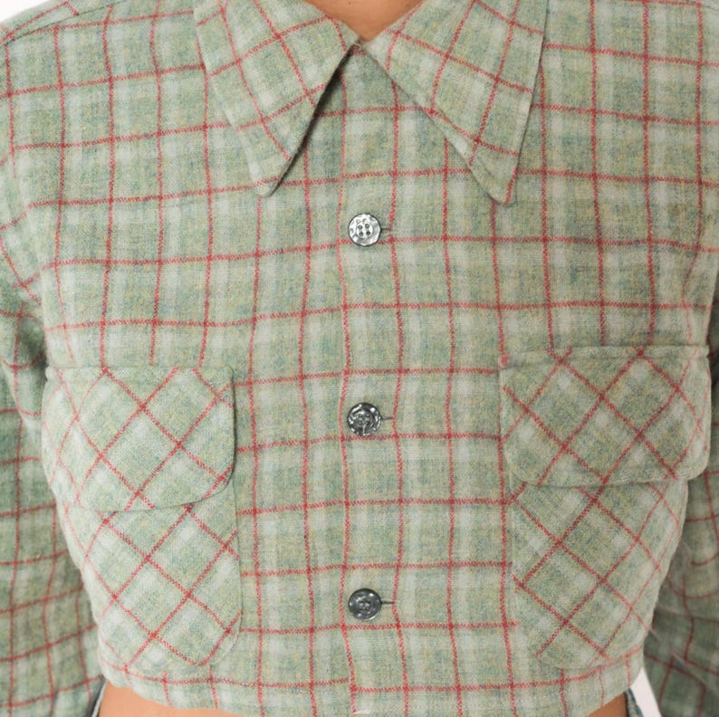 Puede incluir: Una camisa corta abotonada con un patr&oacute;n de cuadros en verde salvia y blanco con finas l&iacute;neas rojas. La prenda presenta un cuello en punta, mangas largas y dos bolsillos delanteros en el pecho con solapas, asegurados por tres botones oscuros.