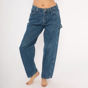 May include: Blue denim carpenter jeans with a relaxed fit. The jeans have a classic five-pocket design, a button and zipper closure, and a hammer loop. The denim is a medium wash, and the jeans appear to be vintage.