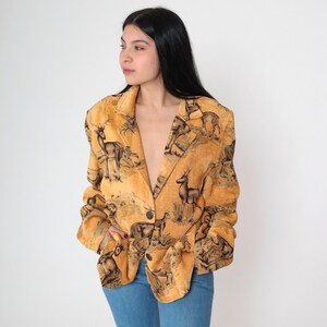 May include: A tan blazer with a wildlife scene print featuring deer and other animals. The blazer has a single-button closure and a classic lapel design. The fabric has a textured appearance, and the overall style is vintage.