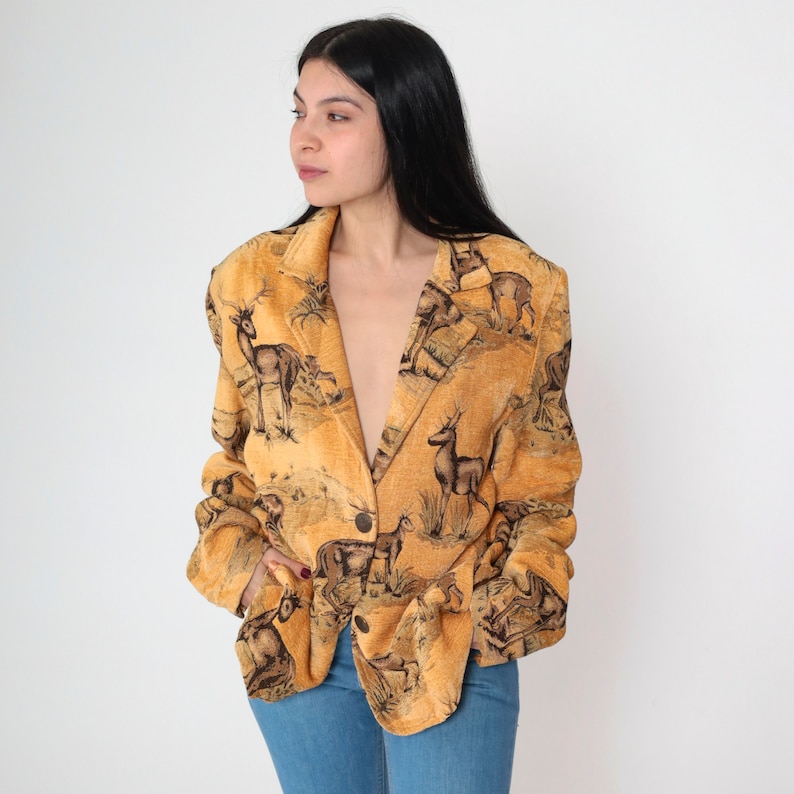 May include: A tan blazer with a wildlife scene print featuring deer and other animals. The blazer has a single-button closure and a classic lapel design. The fabric has a textured appearance, and the overall style is vintage.