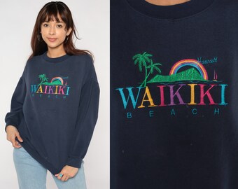Op de afbeelding: Marineblauw sweatshirt met geborduurde tekst "WAIKIKI BEACH" in kleurrijke letters. Het ontwerp bevat een regenboog, palmbomen en het woord "Hawaii". Een casual, comfortabele stijl.