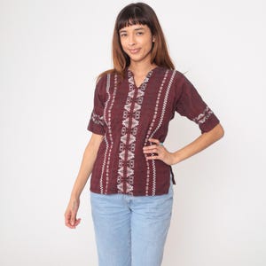 May include: A burgundy short-sleeve shirt with white geometric patterns and vertical stripes. The shirt has a V-neck and is paired with light blue jeans. The sleeves have a decorative white pattern.