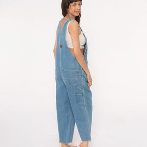May include: Light blue denim overalls with multiple pockets and a frayed hem. The overalls have adjustable straps and a brown patch on the chest. The model is wearing a white top underneath. The overalls are a relaxed fit.