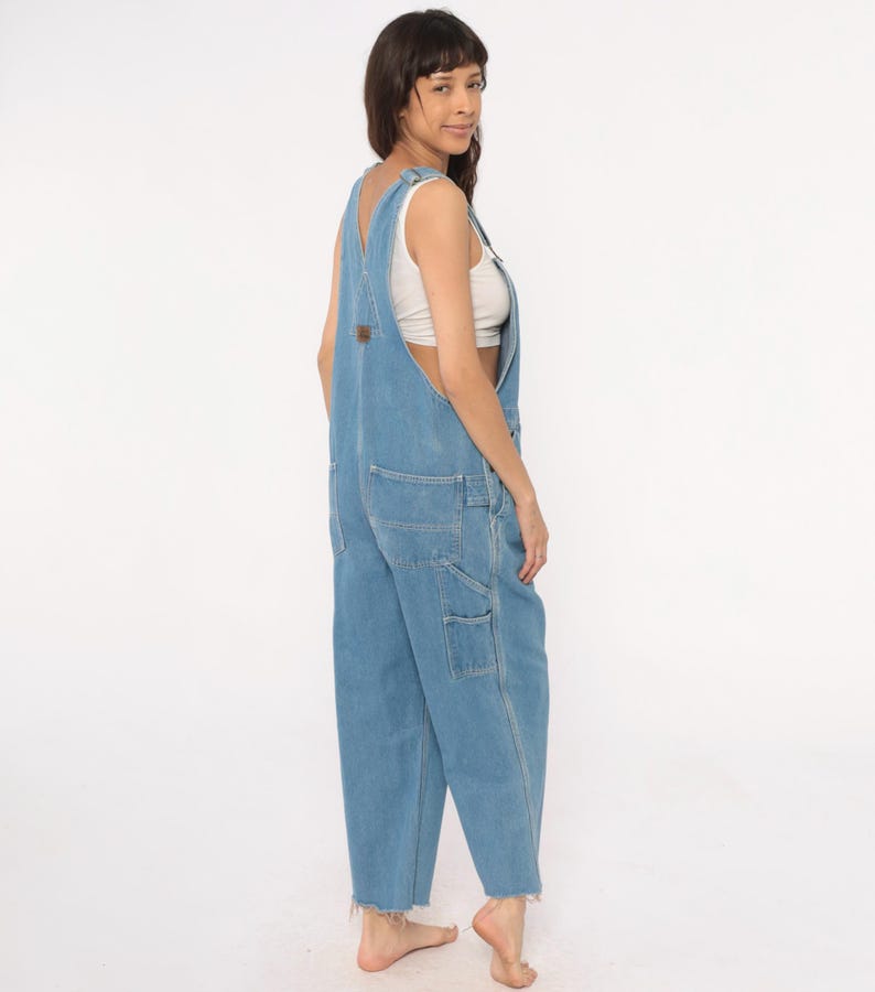 May include: Light blue denim overalls with multiple pockets and a frayed hem. The overalls have adjustable straps and a brown patch on the chest. The model is wearing a white top underneath. The overalls are a relaxed fit.