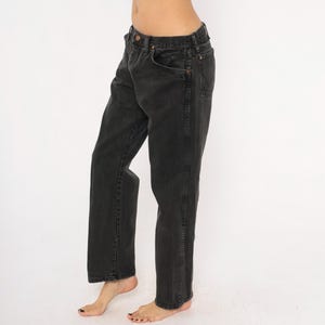May include: Black denim jeans with a classic five-pocket design. The jeans have a straight leg cut and a faded appearance. The image shows the jeans from the waist down, highlighting the details of the fabric and stitching. The jeans are worn.