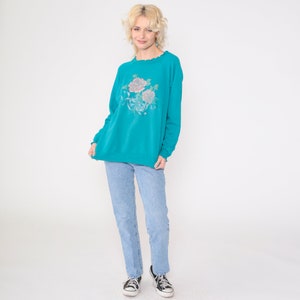 May include: A teal blue sweatshirt with a floral design featuring pink and white roses. The sweatshirt has a ruffled neckline and long sleeves.