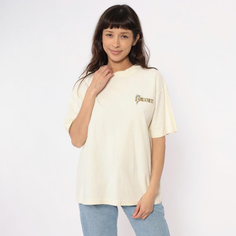 May include: Cream-colored t-shirt with short sleeves. The shirt features the text "GIRLSCOUTS" in a gold-colored script on the upper chest. The model is wearing blue jeans. The shirt has a relaxed fit.