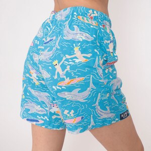 K&ouml;nnte beinhalten: Blaue Badehose mit einem Muster aus Haien, Surfern und Wellen. Die Hose hat ein "Kiwii Surf" Logo an der Seite.