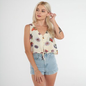 Peut inclure: Un haut sans manches de couleur crème avec un imprimé floral et une fermeture à boutons. Le haut a un nœud à la taille et est court.