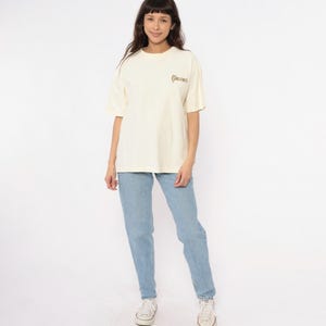 May include: Cream-colored t-shirt with the word "GOLDEN" printed on the upper chest. The shirt is paired with light blue jeans and white sneakers. The model is standing in front of a white background.