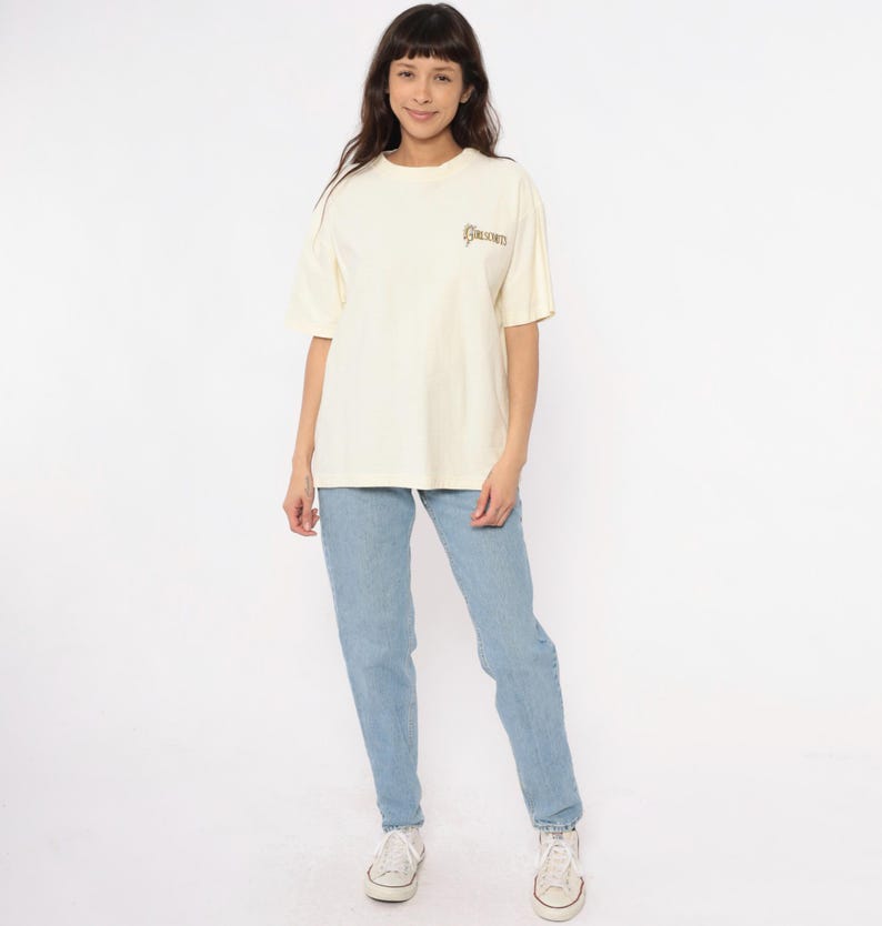 May include: Cream-colored t-shirt with the word "GOLDEN" printed on the upper chest. The shirt is paired with light blue jeans and white sneakers. The model is standing in front of a white background.