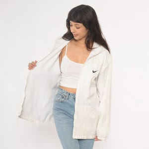 Puede incluir: Chaqueta con capucha blanca Nike con el logotipo negro de Nike en el pecho. La chaqueta est&aacute; abierta, revelando una camiseta sin mangas blanca y vaqueros azul claro. La chaqueta tiene cierre de cremallera.