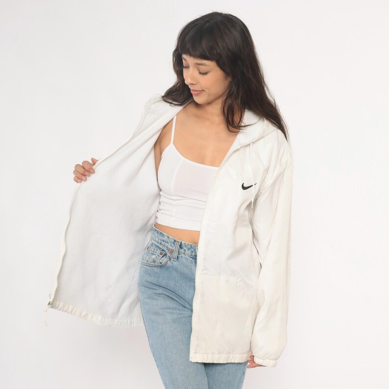 Puede incluir: Chaqueta con capucha blanca Nike con el logotipo negro de Nike en el pecho. La chaqueta est&aacute; abierta, revelando una camiseta sin mangas blanca y vaqueros azul claro. La chaqueta tiene cierre de cremallera.