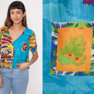 May include: A short-sleeved, V-neck blouse with a bright, colourful print. The top features a patchwork design with a mix of patterns and colours, including blue, yellow, red, and green. The blouse is paired with blue jeans.
