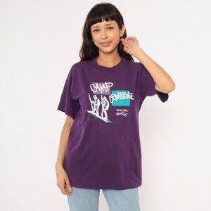 May include: A purple t-shirt with a crew neck featuring the text "CAMP CEDARLEDGE" in white and blue. The graphic shows two figures on a surfboard. The shirt is made of a soft material.